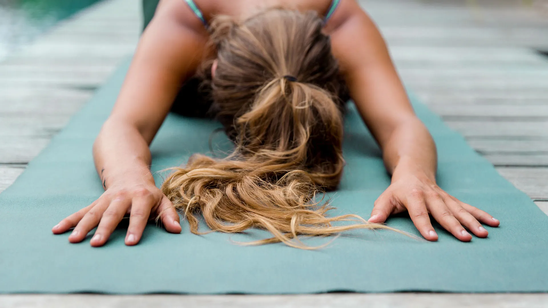 Woman with her hair in a ponytail does a yoga child’s pose outside on a light green yoga mat
